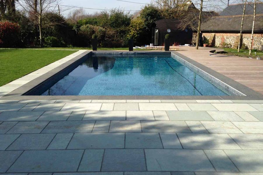 Rectangular swimming pool lined with small tiles, with lawn on 2 sides, decking and Kota Blue limestone paving on the opposite 2.