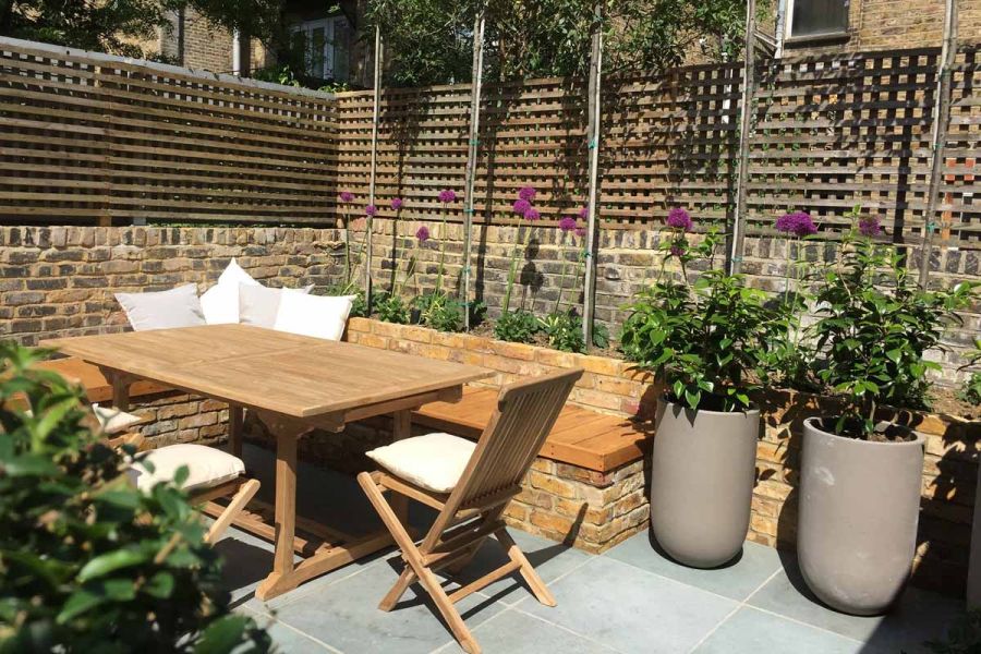Brick-built wood-seated bench with table in corner of urban garden with trellis fixed to top of wall and Kota Blue limestone paving.