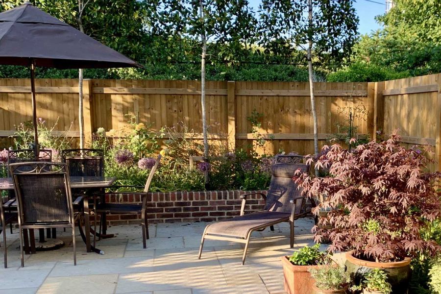 Corner of Kota Brown limestone patio in fenced garden with potted acer, lounger and outdoor dining set.