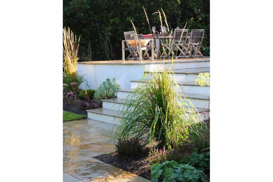5 steps with white risers rise from Kota Brown limestone paving edged with planting to deck area with wooden dining set.