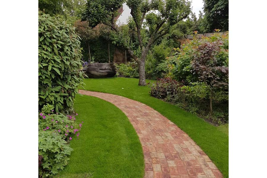 Curved Cotswold Blend clay paver path laid in short-cut grass disappears around bushes. Built by Landscape Artisan.