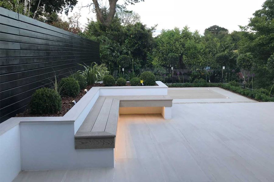 Very large area paved with Faro Porcelain patio tiles. Edged with L-shaped raised bed with formal planting and wood-topped bench.