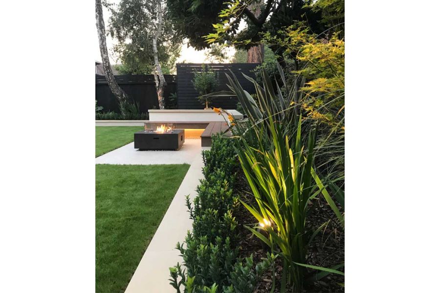 Lush planting overhangs Golden Stone porcelain paving path to patio with firepit and underlit bench. By Landscape Design Studio.