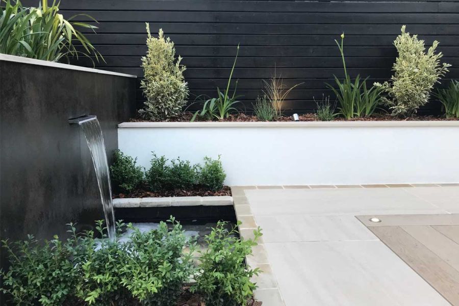 Water feature cascades into pool below from wall faced in Steel Dark Luxury DesignClad, by paved area with planted raised bed.