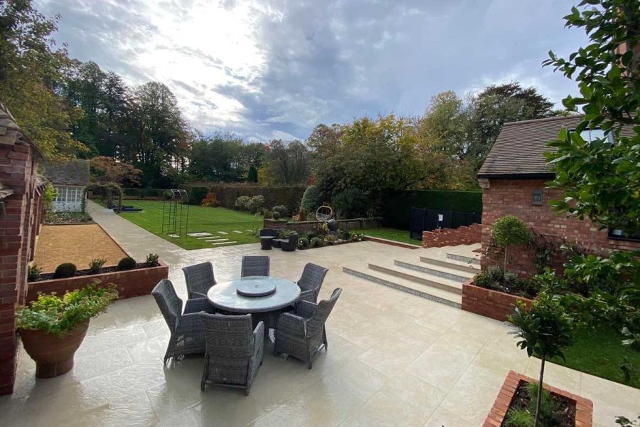 Large patio in Golden Stone porcelain paving with path leading past lawn and garden building. Design by Wyre Hall Landscapes.
