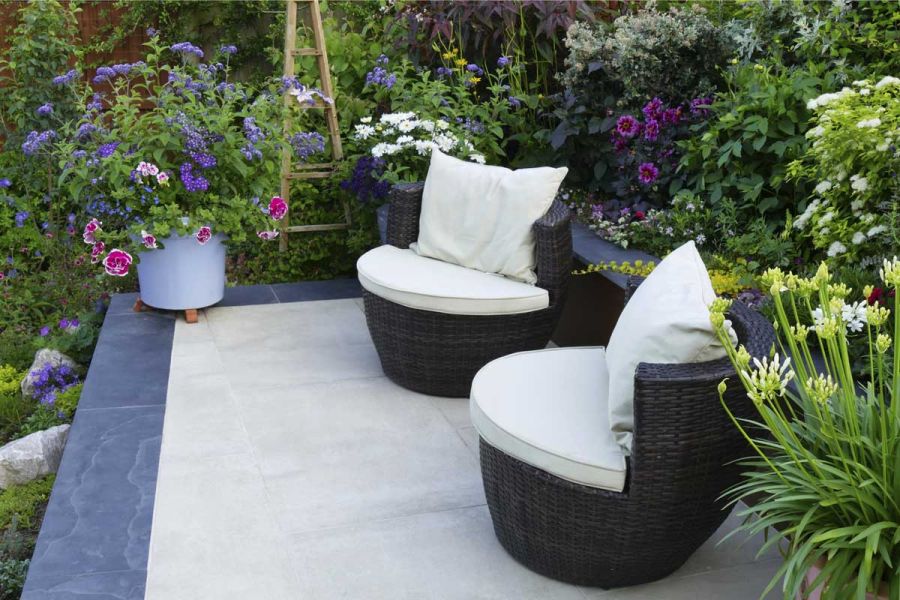 2 rattan barrel chairs on a small raised patio made from Cream porcelain edged with dark porcelain surrounded by vibrant summer planting.