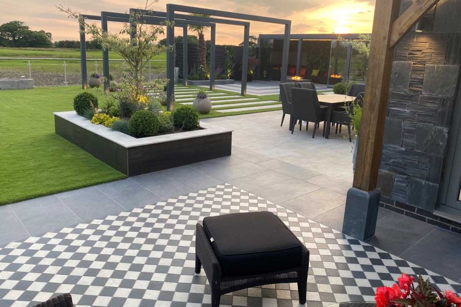 Stool sits on chequerboard paving of light grey and charcoal porcelain setts leading to large patio with pergola and garden room.