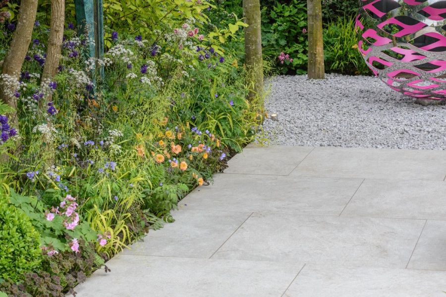 Silver-grey porcelain paving leading to white gravel with spherical pink and silver metal sculpture, edged by lush flower border.