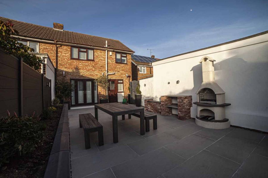 At rear of house, a wooden table and benches stand on urban grey porcelain paving near white chiminea, raised bed on left.