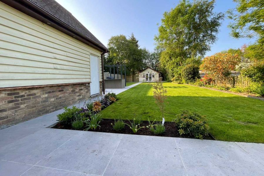 Small porcelain patio adjoining a long lawn with mature shrubs and trees down one side and leading towards a timber clad summer house.