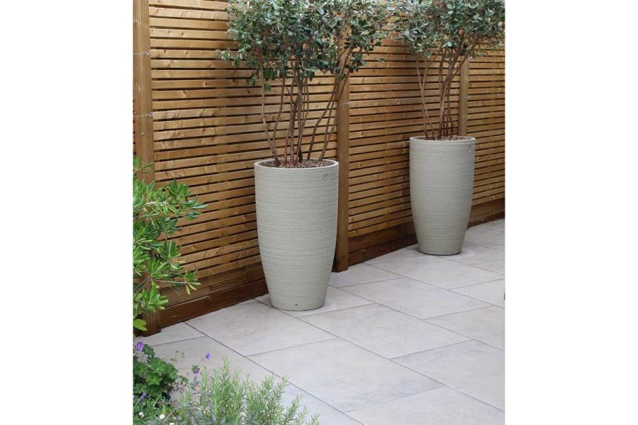 Jura Grey Porcelain paving used alongside wooden fencing, two tall grey pots stand on the paved area, with small trees planted.