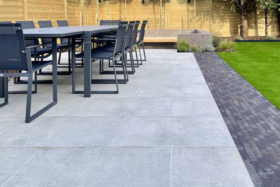 Long metal outdoor dining set on top of a porcelain paving patio edged with dark Clay Pavers and an artificial lawn.