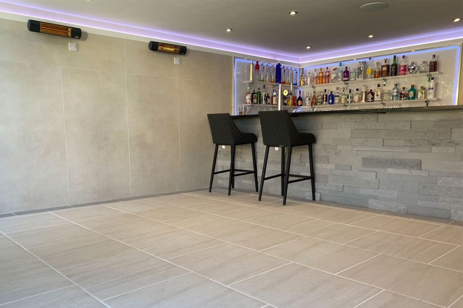 Bar paved in Urban Grey Porcelain with pale grouting. Two black stools at stone-clad counter. Built by Limebok Landscaping.