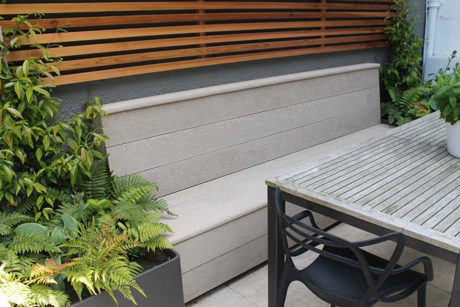 Skirted bench constructed of Limed Oak Millboard decking planks sits under slatted fencing, next to planter with ferns.