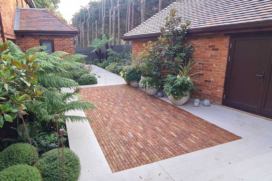 Rectangle of Cotswold Blend Clay Paver Bricks laid into pale paving between brick buildings. Bed with box balls and foliage to left.