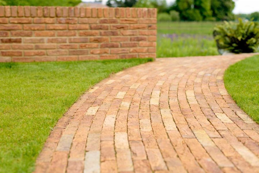 Curved path of Cotswold Blend clay pavers, laid lengthways in direction of travel, leads across lawn, past wall to grassy area.