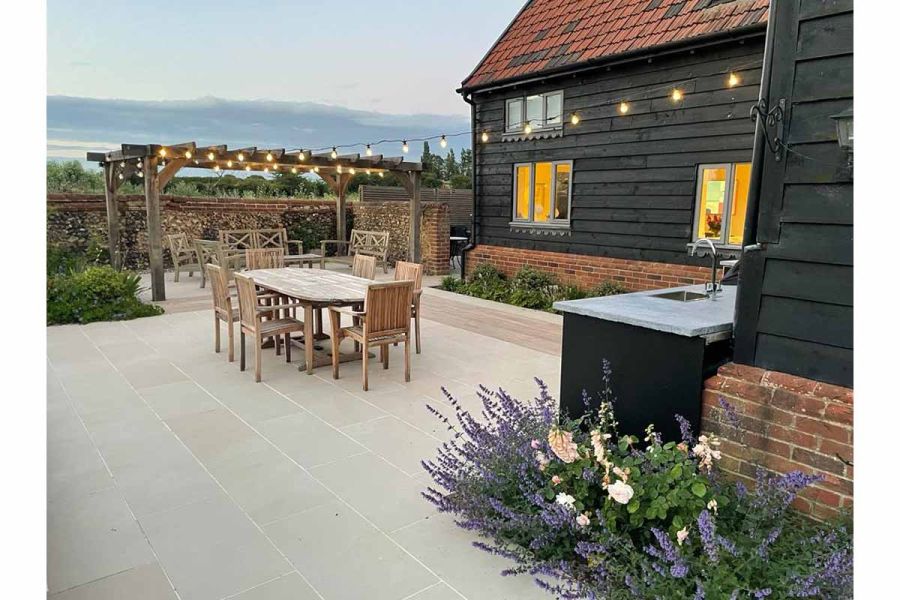 string lights from house to wooden pergola with dining underneath, slab khaki porcelain paving shown at dusk with country views.
