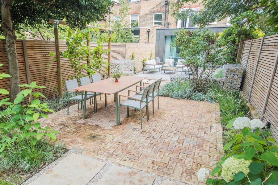 London Mixture Clay Paver Bricks alongside a wooden slatted fence. A patio with a table for six around lush greenery in the background.