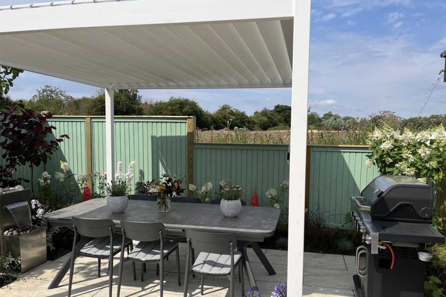 Green fence behind Proteus white aluminium pergola, with closed roof, covering dining set between gas barbecue and water feature.