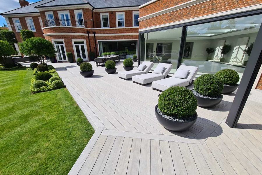 Long, wide, angled patio of Luna composite decking with 3 sunloungers, potted shrubs and lawn with topiary. Built by Stone & Webb.