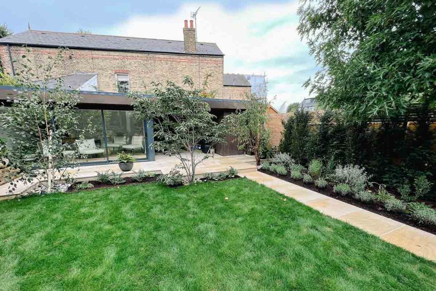 Straight path of Harvest sawn sandstone leads between lawn and planted border to raised patio of house with glass sliding doors.