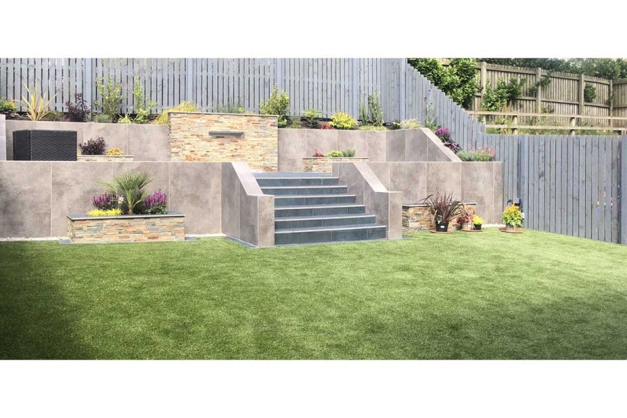 Artificial lawn laid in front of a large raised terrace featuring a central set of dark porcelain steps and raised flower beds.