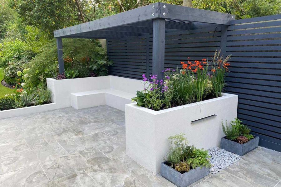 Marble Grey porcelain garden tiles UK pave pergola shaded seating area, with wall-mounted water feature and colourful planting.