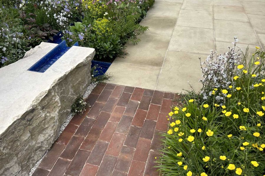 Dorset Antique Belgian bricks laid between low wall with rugged white coping and yellow-flowered shrub, next to stone paving.