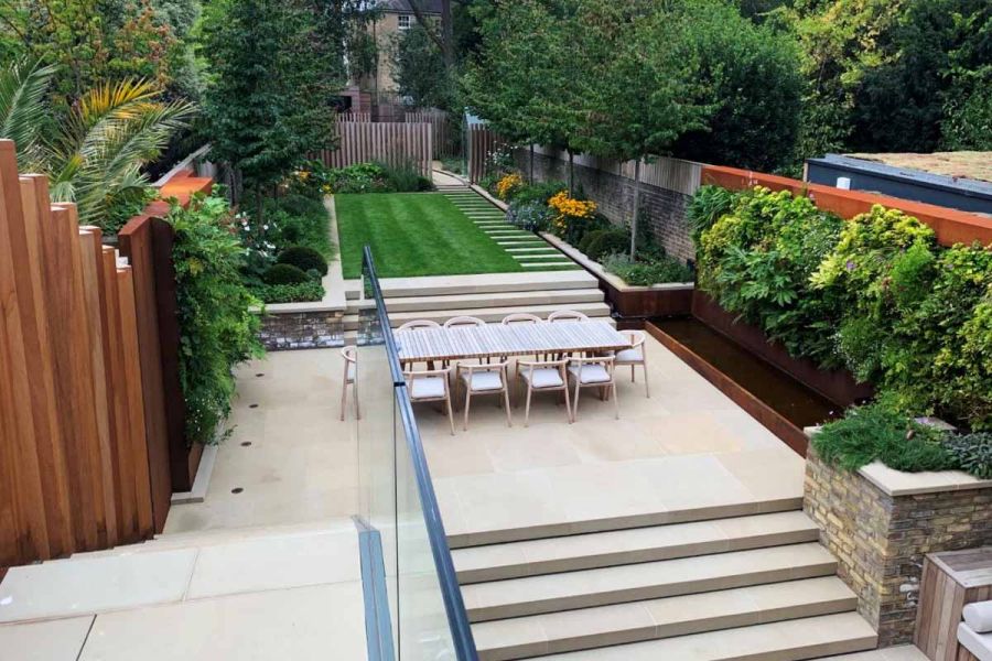 Steps with glass railings descend to long, narrow garden with borders down each side and large Britannia Buff sawn Yorkstone patio.