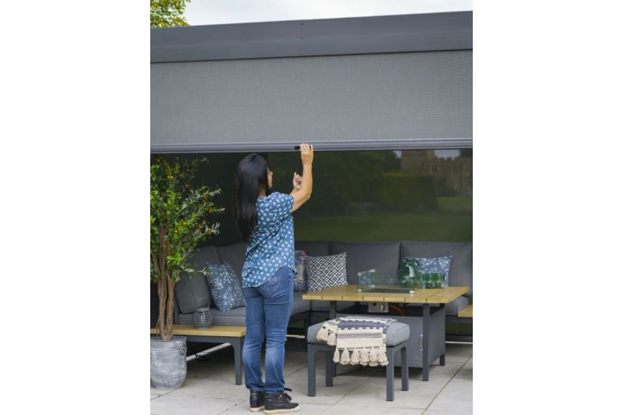 Woman pulls down one of adjustable sides of Dark Grey Metal Pergola, which stands over table and modular sofa.