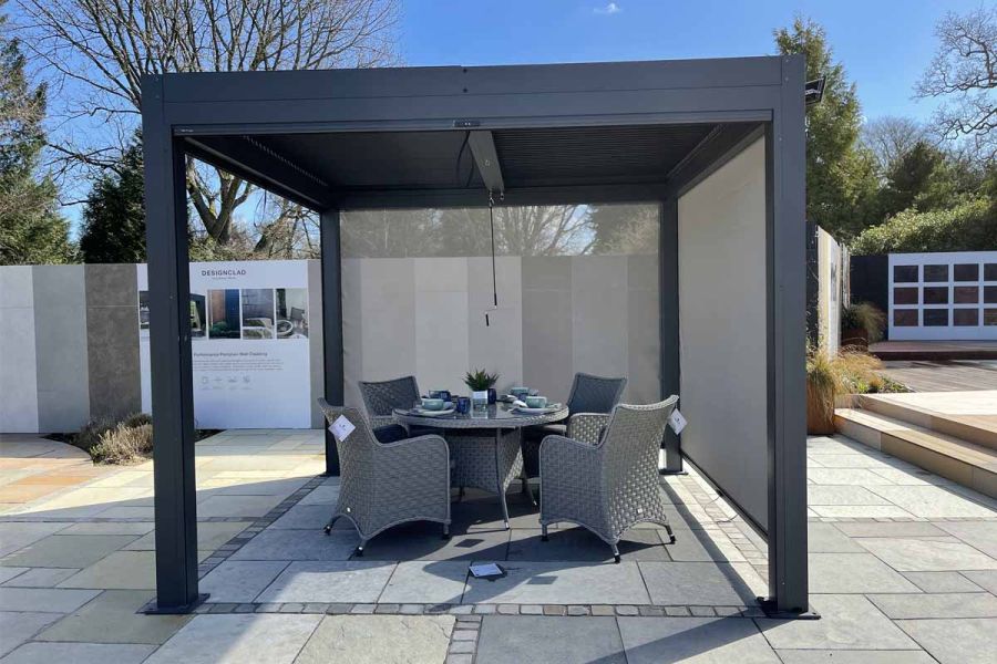 LG Dark Grey Metal Pergola, 2 side blinds lowered, over rattan modern garden furniture standing on natural stone paving.