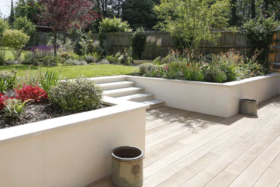 White-walled terraced beds on either side of 5 wide steps from lawn surrounded by beds to Golden Oak Millboard composite decking.