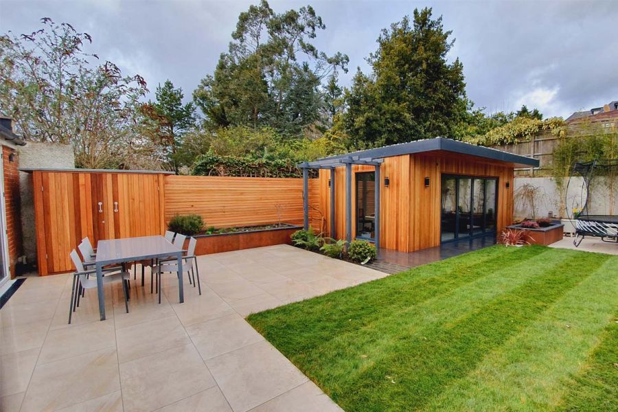 Porcelain patio with grey dining table and chairs, recently laid lawn, cedar screens and a bespoke cedar clad garden building.