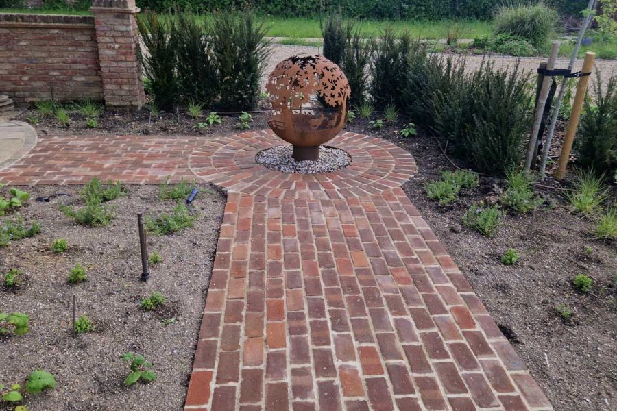 Pierced fire bowl sits in paved circle at right angle of path of Dorset Antique Belgian bricks between beds with young plants.