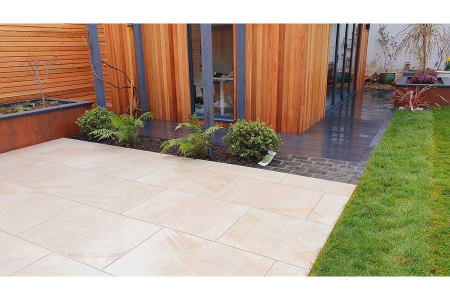 Ash Beige porcelain patio in front of a wooden cladded garden building, a raised flower bed on one side and a lawn on the other side.