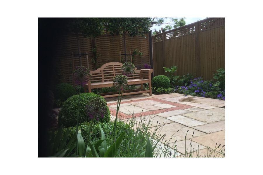 Lutyens-style bench at one end of Mint Indian sandstone paving with brick detail, next to border below tall fence.