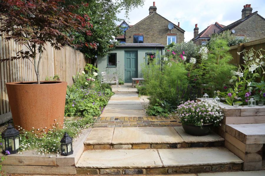 2 shallow steps with brick risers ascend to straight path of Mint Indian sandstone slabs leading to garden shed. 