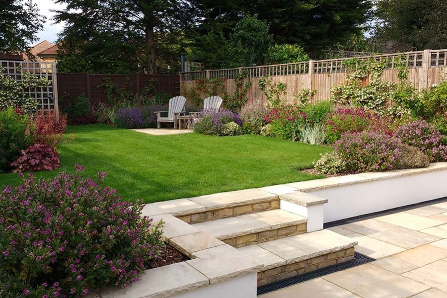 White retaining wall with 3 steps rising from Mint Indian sandstone patio to lawn with planted borders in small suburban garden.