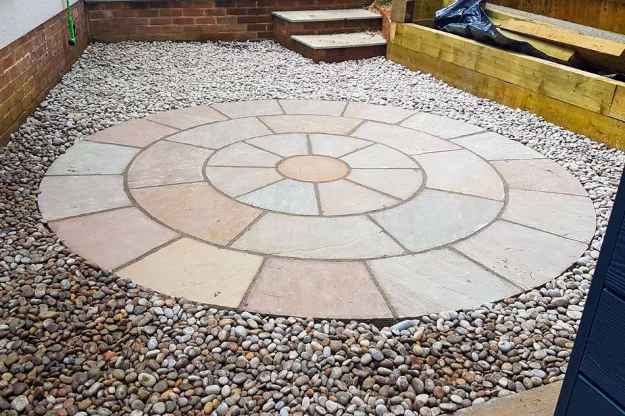 Gravelled area bordered by wall and sleepers, with Mint sandstone paving circle. 2 steps in far right corner. Design by Silva Tree.