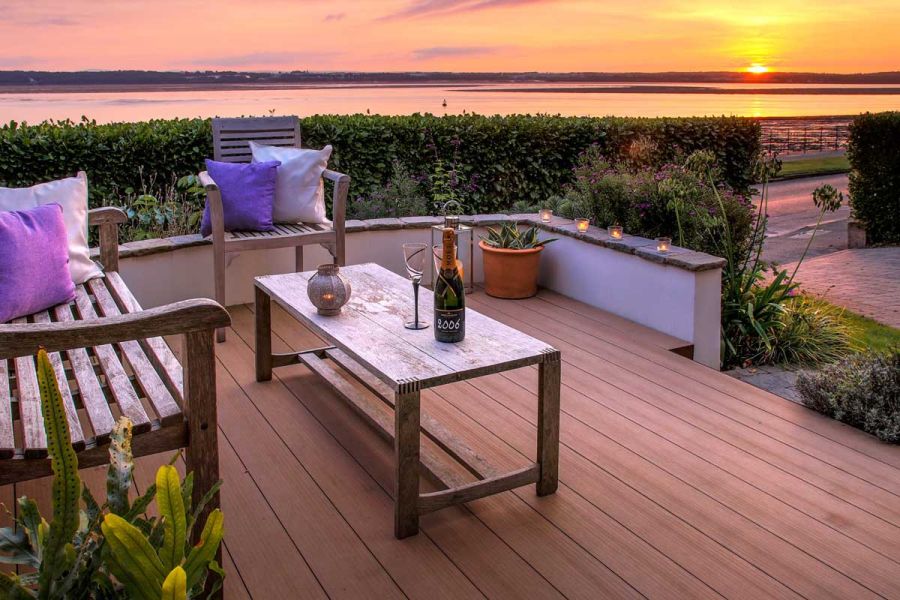 Low, white-painted wall surrounds front garden laid with Mocha WPC decking, with bench, table and wide view over water to sunset.