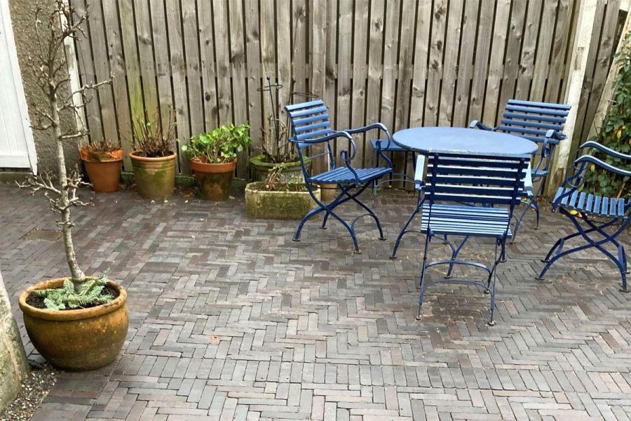 Blue garden set sits near corner of house on Moderna clay pavers laid herringbone next to fence with range of planters. 