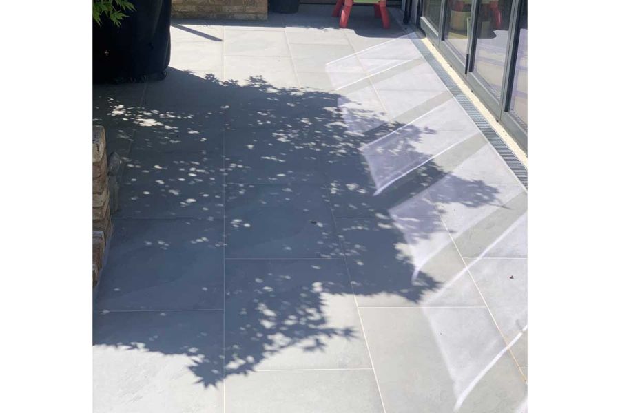 Light reflects from bifold doors onto Florence Storm porcelain paving with dark tree shadow. Laid by Monarch Lawn and Landscape.