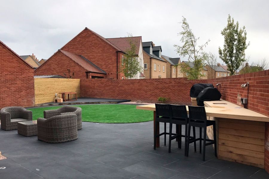 Florence Dark porcelain paves walled garden with bar and stools, rattan furniture and central lawn, by Muddy Boots Landscaping.