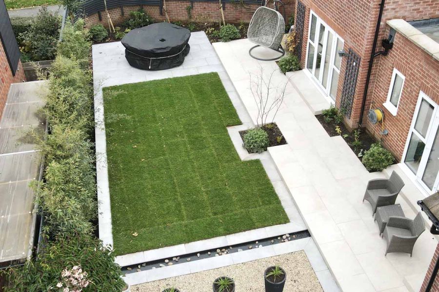 View down on rectangular lawn framed with Florence Grey paving, next to beige patio running along back of house. 