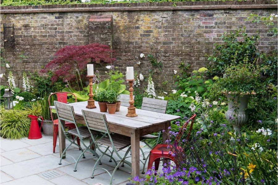 Wooden dining set on Kota Blue limestone paving edged by verdant, flowery planting , with tall brick boundary wall.