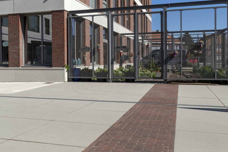 Wide strip of Novara clay pavers, set into large format cream paving, leads to glassed-in planted area next to office building.