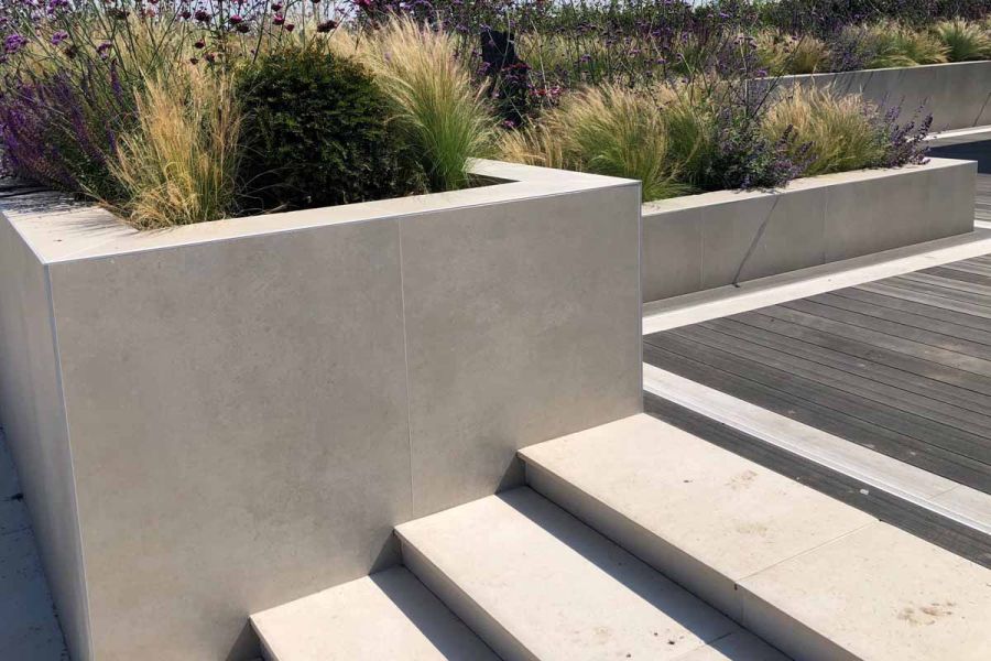 Raised beds of different heights flank composite decking and porcelain steps, clad in Off White exterior cladding with pale grout.