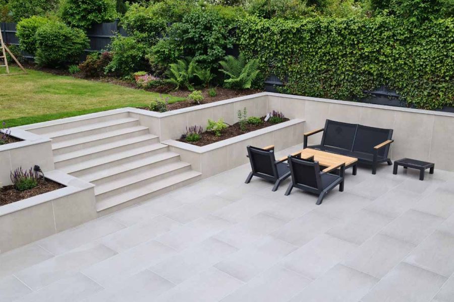 Porcelain paving with black luxury garden furniture, retaining walls in Off White exterior cladding, and wide steps up to lawn.