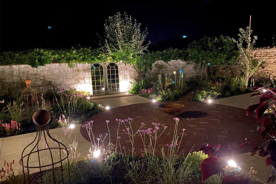 Pale-walled garden at night. Wide white paths separate 4 large beds, around large central circle of Old English Clay Pavers.