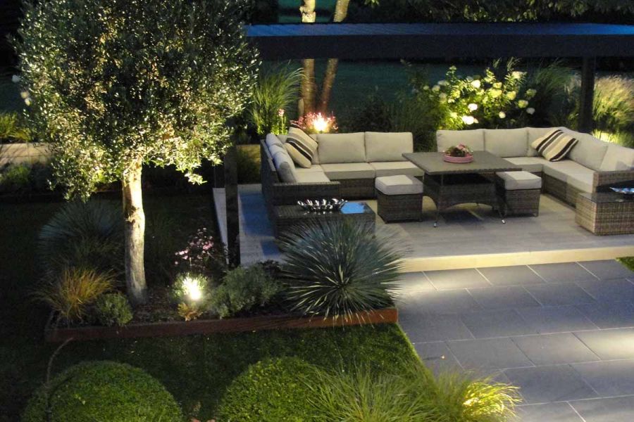 A nighttime view shows densely planted garden with an illuminated pergola, under which lies rattan furniture, all set atop silver Contro porcelain paving.